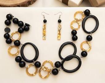 May include: A jewelry set consisting of a multi-strand necklace and matching drop earrings. The necklace features black spherical beads, large black oval rings, and textured gold-tone circular links. The earrings have a black bead and a gold-tone bar.