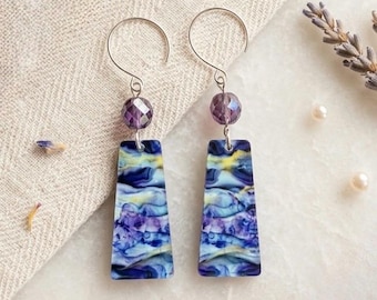 May include: A pair of dangle earrings featuring silver-toned wire hooks, each suspending a single faceted purple bead above a flat, trapezoidal pendant. The pendants display a marbled pattern of swirling blue, purple, and yellow hues.