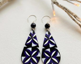 May include: A pair of teardrop-shaped dangle earrings featuring a geometric pattern in black, white, and deep purple. Each earring is suspended from a silver-toned hook and accented with a single faceted black bead at the top of the pendant.