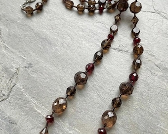 May include: A long necklace featuring faceted translucent brown beads and smaller deep red beads arranged in a repeating pattern. The necklace is finished with a silver-toned metal toggle clasp, creating a continuous strand of alternating dark brown and red glass-like elements.