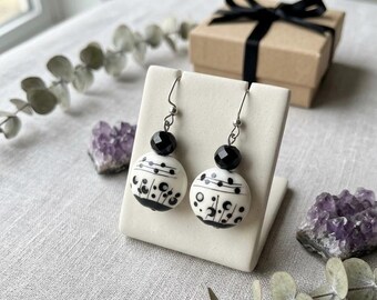 May include: A pair of drop earrings featuring round white ceramic beads with black hand-painted line and dot patterns. Each earring includes a smaller faceted black bead above the ceramic sphere, suspended from a silver-toned metal hook wire.