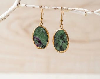 May include: A pair of oval-shaped earrings featuring natural green and magenta stone slices with gold-toned edges. The stones are suspended from gold-toned metal hooks, showcasing a raw, textured surface with distinct mineral inclusions.