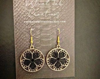 May include: A pair of gold-colored earrings with a circular design. Each earring features a black, flower-shaped center within a gold filigree frame. The earrings are displayed on a textured card with the text "Virtual Tab Creations" and the Etsy shop URL.