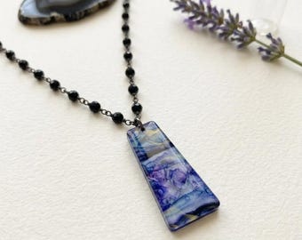 May include: A necklace featuring a long, trapezoidal pendant with a marbled blue, purple, and white pattern. The pendant hangs from a delicate chain composed of small, faceted black beads connected by thin dark metal links.