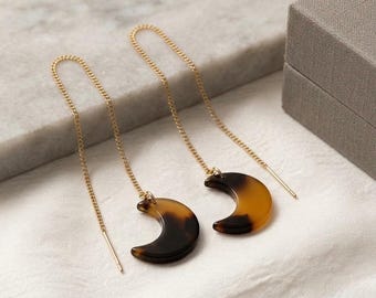 May include: A pair of gold-tone chain threader earrings featuring crescent moon-shaped charms. The charms are made of a polished, translucent material with a mottled brown and black tortoiseshell pattern, suspended from delicate gold-tone metal chains.