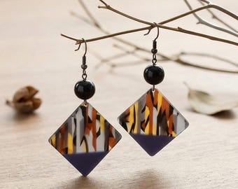May include: A pair of dangle earrings featuring a black spherical bead attached to a diamond-shaped pendant. The pendant has a top section with a multi-colored pattern of white, orange, brown, and gray, and a solid purple triangle at the bottom.