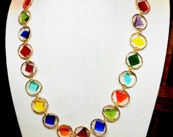 May include: A necklace featuring a series of small, multi-colored geometric glass shapes, including squares and triangles, each encased in a thin gold-toned metal ring. The glass pieces are arranged in a sequence of red, green, blue, and yellow.