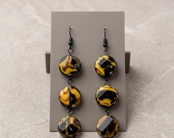 May include: A pair of dangle earrings featuring three circular beads each, showcasing a marbled pattern of black and golden yellow. The beads are connected by black metal jump rings and hang from black metal fishhook ear wires on a gray card.