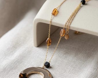 May include: A gold-tone chain necklace featuring a large circular pendant with a split design of polished gold and brown tortoiseshell pattern. The chain is accented with small amber-colored translucent beads and a single black cube bead.