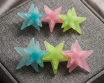May include: Two hair clips, each featuring three layered star-shaped charms in translucent glittery finishes. The top clip has blue, pink, and lime green stars, while the bottom clip features blue, lime green, and pink stars arranged horizontally.