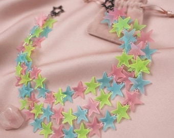 May include: A multi-layered necklace featuring numerous translucent star-shaped charms in shades of neon green, light blue, and soft pink. The stars are linked together in a cascading pattern, finished with a black star-shaped clasp closure.