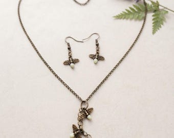 May include: A jewelry set consisting of a necklace and a pair of earrings. The necklace features a bronze-toned chain with a pendant of three small bee charms, each with a pale, off-white bead body. The matching earrings are bee-shaped dangles.