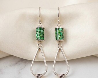 May include: A pair of silver-toned drop earrings featuring a cylindrical bead filled with green mosaic-style inlay. Below the cylinder hangs a teardrop-shaped silver frame with a small, textured, rope-like detail at the bottom point.