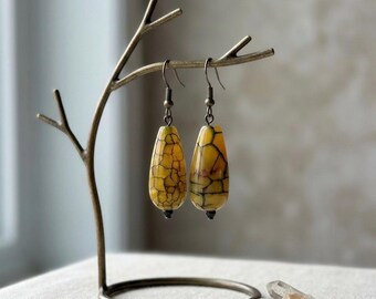 May include: A pair of teardrop-shaped earrings featuring a yellow stone with a dark brown crackle pattern. The earrings hang from bronze-toned metal hooks and are suspended from a bronze-colored metal display stand shaped like a tree branch.