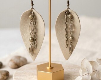 May include: A pair of cream-colored leather leaf-shaped earrings hanging from a gold-toned T-bar stand. Each earring features a bronze-colored metal chain, two metallic beads, and two translucent teardrop-shaped beads suspended from the leather.