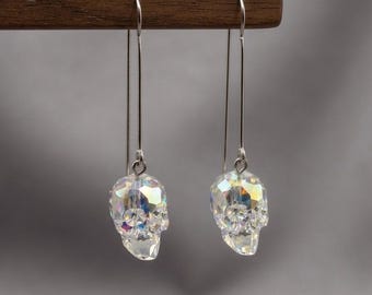 May include: A pair of drop earrings featuring faceted, iridescent crystal skulls suspended from long, slender silver-toned metal hooks. The skulls display a multi-colored, reflective sheen, catching light to reveal subtle rainbow hues against the clear glass.
