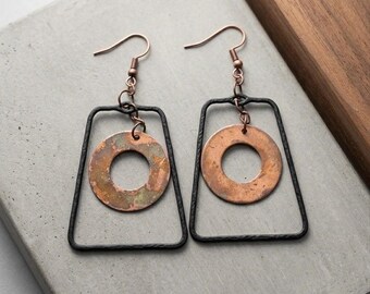 May include: A pair of dangle earrings featuring a black metal trapezoidal frame with a textured surface. Inside each frame hangs a circular copper disc with a weathered, patina finish. The earrings are attached to copper-colored metal ear wires.