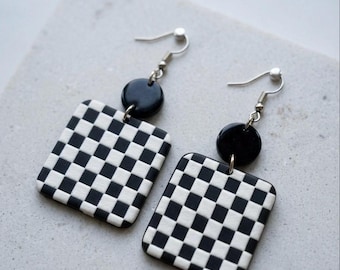 May include: A pair of dangle earrings featuring a black and white checkered square pendant suspended from a small, solid black circular stud. The earrings are attached to silver-toned metal hooks, creating a geometric design with high contrast.