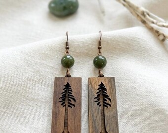 May include: A pair of rectangular wooden drop earrings featuring a laser-cut pine tree silhouette. Each earring is accented with a single round, polished dark green bead and suspended from a bronze-toned metal hook wire finding.