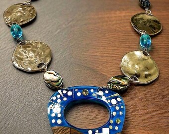 May include: A necklace featuring a large, teardrop-shaped pendant with a blue enamel top, white dot accents, and a wood-grain base with inlaid white architectural shapes. The chain consists of hammered gold-tone discs, blue glass beads, and iridescent abalone shell beads.