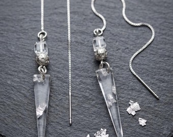 May include: A pair of silver-toned threader earrings featuring long, clear, triangular resin pendants embedded with silver leaf flakes. Each earring includes a small clear cube bead and a textured silver-toned spherical bead above the pendant.