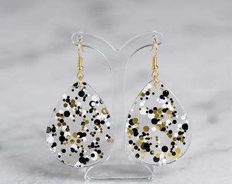 May include: A pair of teardrop-shaped earrings made of clear acrylic, featuring an embedded confetti pattern of black, white, and metallic gold circular dots. The earrings hang from gold-toned metal fishhook wires on a clear display stand.