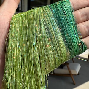 May include: A bundle of shimmering green craft thread, transitioning from light chartreuse to a darker green. The threads have a metallic, glittery appearance. The strands are long and flowing, ideal for various craft projects.