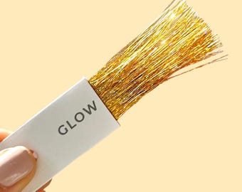 May include: A rectangular white tag with the word GLOW printed in black, attached to a bundle of thin, metallic gold tinsel strands. The text LIGHT GOLD is printed in the bottom right corner of the image against a pale yellow background.