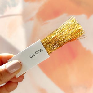 Puede incluir: Una pequeña tarjeta blanca con la palabra "GLOW" impresa en ella. Un manojo de hebras de purpurina dorada está unido a la tarjeta.