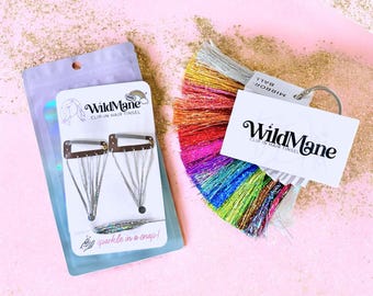 May include: WildMane clip-in hair tinsel set featuring two silver hair clips on a branded card and a multi-colored fan of tinsel strands attached to a ring with a WildMane tag. The packaging includes the text WildMane Clip-in Hair Tinsel and Sparkle in a snap!