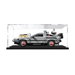 May include: A grey brick-built model car featuring a tan hood component, black wheels with red centers, and rear mechanical details, all enclosed within a clear rectangular acrylic display case with a solid black base for protection and viewing.