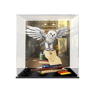 May include: A white brick-built owl with spread wings perched on a stack of books, inside a clear display case. The display includes a brown wand, a letter with a wax seal, a small green bottle, a golden snitch, and a set of red and yellow bricks.