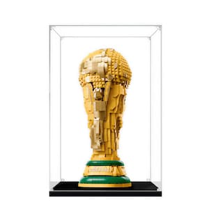 May include: A gold-colored trophy constructed from interlocking plastic building bricks, featuring a spherical top and a green base with the text FIFA WORLD CUP printed on it, all enclosed within a clear rectangular acrylic display case.