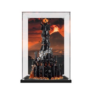 May include: A black brick-built tower model featuring a flaming eye at the peak, enclosed within a clear rectangular acrylic display case. The background inside the case depicts a volcanic landscape with orange and red hues, mimicking lava.
