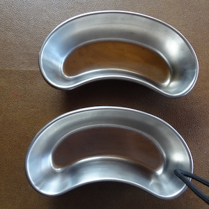 May include: Two stainless steel kidney dishes, one on top of the other. The top dish is slightly tilted to the right. The bottom dish has a black elastic band attached to the side.