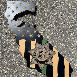 May include: A wooden cutout of the state of California with an American flag design in the top portion and a black and tan striped design in the bottom portion. A green and gold star with the text "DEPT. OF CORRECTIONS & REHABILITATION" is centered on the striped portion.