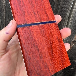 Op de afbeelding: Een rechthoekig houten blok met een rijke, roodbruine tint. Een dunne, donkerblauwe inlegstrip verdeelt het hout, wat een contrasterend detail toevoegt. De houtnerf is prominent aanwezig en de randen zijn schoon afgewerkt.