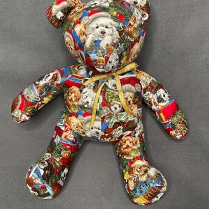 May include: A stuffed teddy bear made of fabric featuring a repeating pattern of various dog breeds, Christmas trees, and red ornaments. The bear has a gold ribbon tied around its neck and displays a colorful holiday-themed print throughout.