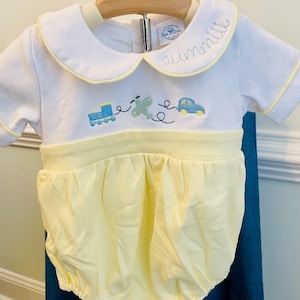 May include: White and yellow baby romper with a Peter Pan collar. The romper features embroidered images of a train, airplane, and car. The collar is trimmed in yellow and has the word "summit" embroidered on it.