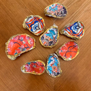 May include: Nine oyster shells, each covered in a different patterned paper, with gold accents. The patterns are floral, geometric, and abstract, in shades of red, blue, and gold.