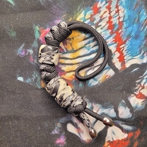 May include: A black and grey paracord lanyard featuring a marbled white and black bead. The lanyard has a loop and two silver-coloured metal end caps. The background is a colourful tie-dye pattern.