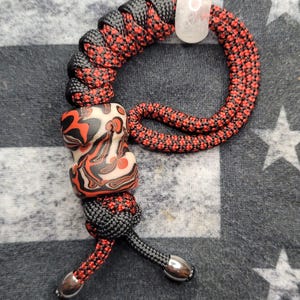 May include: A handmade lanyard with a red and black woven cord. It features a decorative bead with red, black, and white patterns, a clear crystal bead, and silver-toned metal end caps.