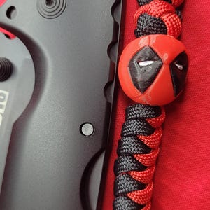 K&ouml;nnte beinhalten: Schwarzes Klappmesser mit einem rot-schwarzen Schl&uuml;sselband mit einer Deadpool-Perle. Das Schl&uuml;sselband besteht aus geflochtenem Kordel und hat eine schwarze Perle am Ende. Die Deadpool-Perle ist rot mit schwarzen Akzenten.