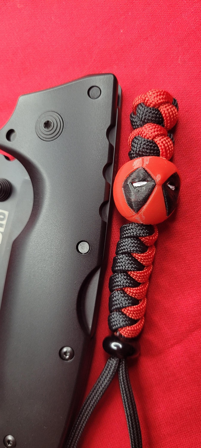 K&ouml;nnte beinhalten: Schwarzes Klappmesser mit einem rot-schwarzen Schl&uuml;sselband mit einer Deadpool-Perle. Das Schl&uuml;sselband besteht aus geflochtenem Kordel und hat eine schwarze Perle am Ende. Die Deadpool-Perle ist rot mit schwarzen Akzenten.