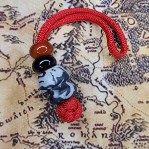 Op de afbeelding: Een rood koord met een knoop aan het einde en vier kralen. De kralen zijn rood, zwart en grijs met een marmerpatroon. Het koord ligt op een stoffen achtergrond met een kaart erop gedrukt.