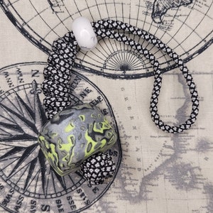 Puede incluir: Un cordón trenzado blanco y negro con una cuenta blanca y una cuenta con estampado verde y gris. El cordón está sobre un fondo de tela con un diseño de mapa.