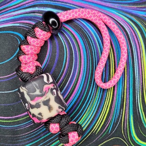 May include: A handmade lanyard with a black and pink braided cord and a decorative bead. The bead has a cream base with black and pink abstract patterns. The lanyard also has a pink cord loop and a black bead.