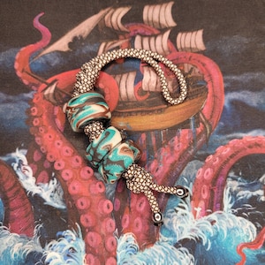 May include: A teal, brown, and white patterned beaded lanyard with silver metal accents. The lanyard is on a background of an octopus and a ship.