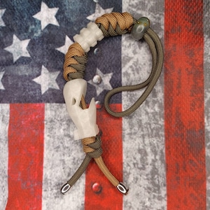 May include: A white paracord bracelet with a white skull-shaped charm and a green and brown paracord strap. The bracelet is on a red, white, and blue American flag background.