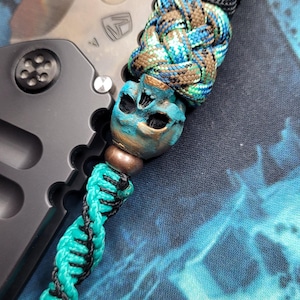 Puede incluir: Un cordón de cuchillo personalizado con una cuenta de calavera y un cordón trenzado. La cuenta de calavera está pintada con tonos azules y bronce. El cordón presenta un cordón trenzado negro y verde azulado y una cuenta marrón. La hoja del cuchillo es visible en el fondo.